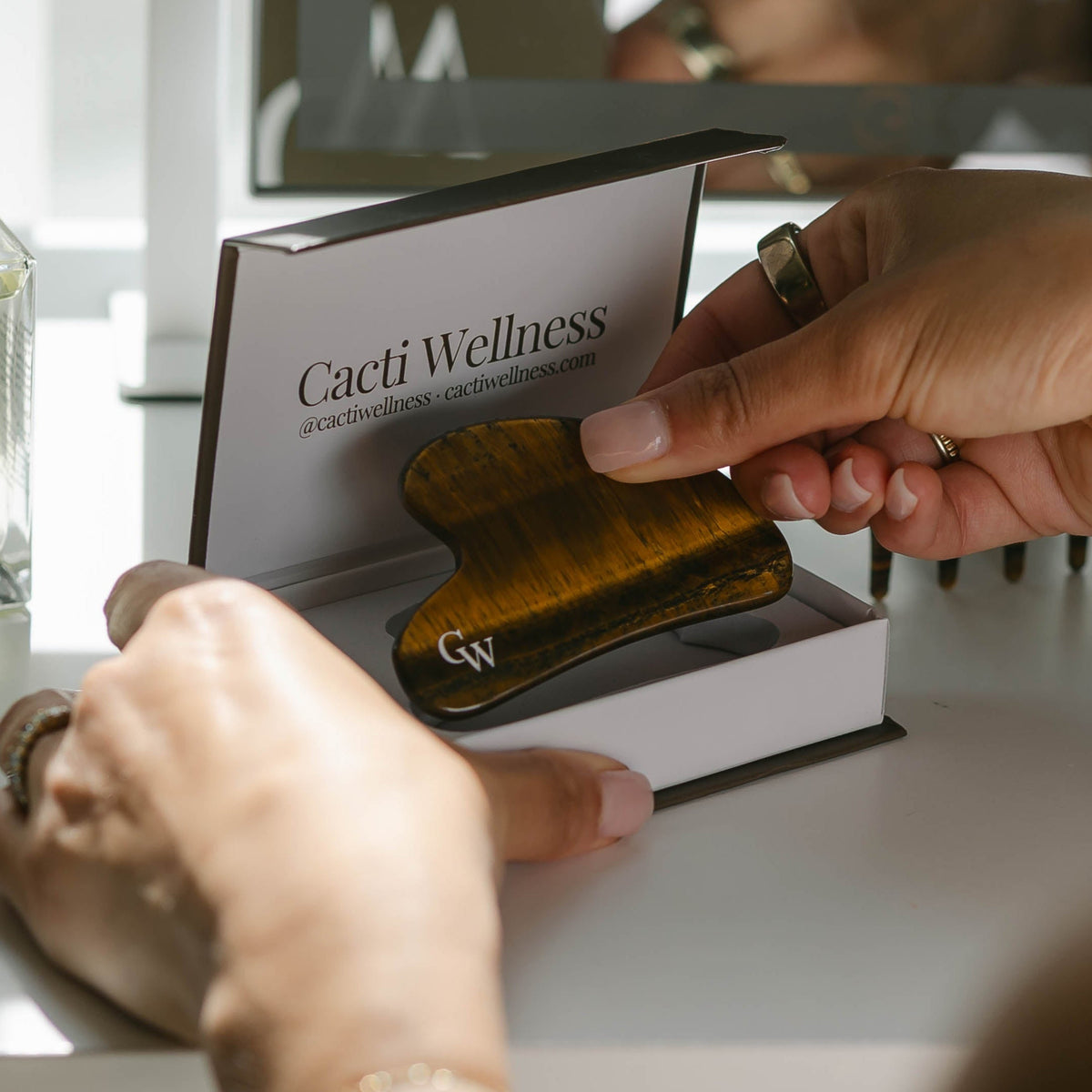 Person holding The Gua Sha by Cacti Wellness on a vanity with skincare products